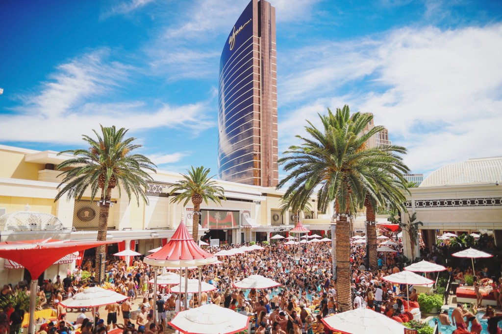 Photo showing a crowd partying at Encore Beach Club at the Wynn in Las Vegas. Blue skies and palm trees line the resort, with the Wynn hotel sticking straight up in the background.